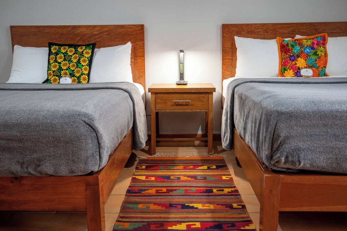 A hotel room with two beds, each with a wooden headboard, a nightstand between them, and a colorful rug on the floor.