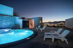 A rooftop pool area with a hot tub, lounge chairs, and a view of a cityscape at dusk.