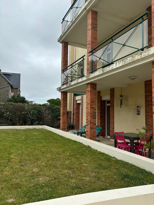Un edificio de dos pisos con un balcón, columnas de ladrillo y un pequeño jardín con muebles para exteriores.