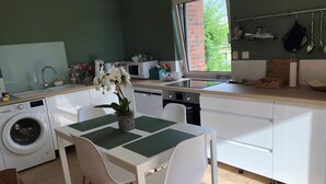A modern kitchen with a built-in washing machine, a dining area with a table and chairs, and a window providing a view of greenery outside. - Granville, 50m from the sea, on the garden level, nice and cosy apartment-terrace for 2 persons (Granville)