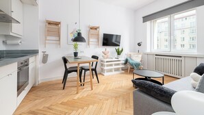 Un appartement moderne comprenant une cuisine, un coin repas et un salon. Il est doté d’une grande fenêtre, d’un téléviseur à écran plat et d’un parquet en bois disposé en point de Hongrie.