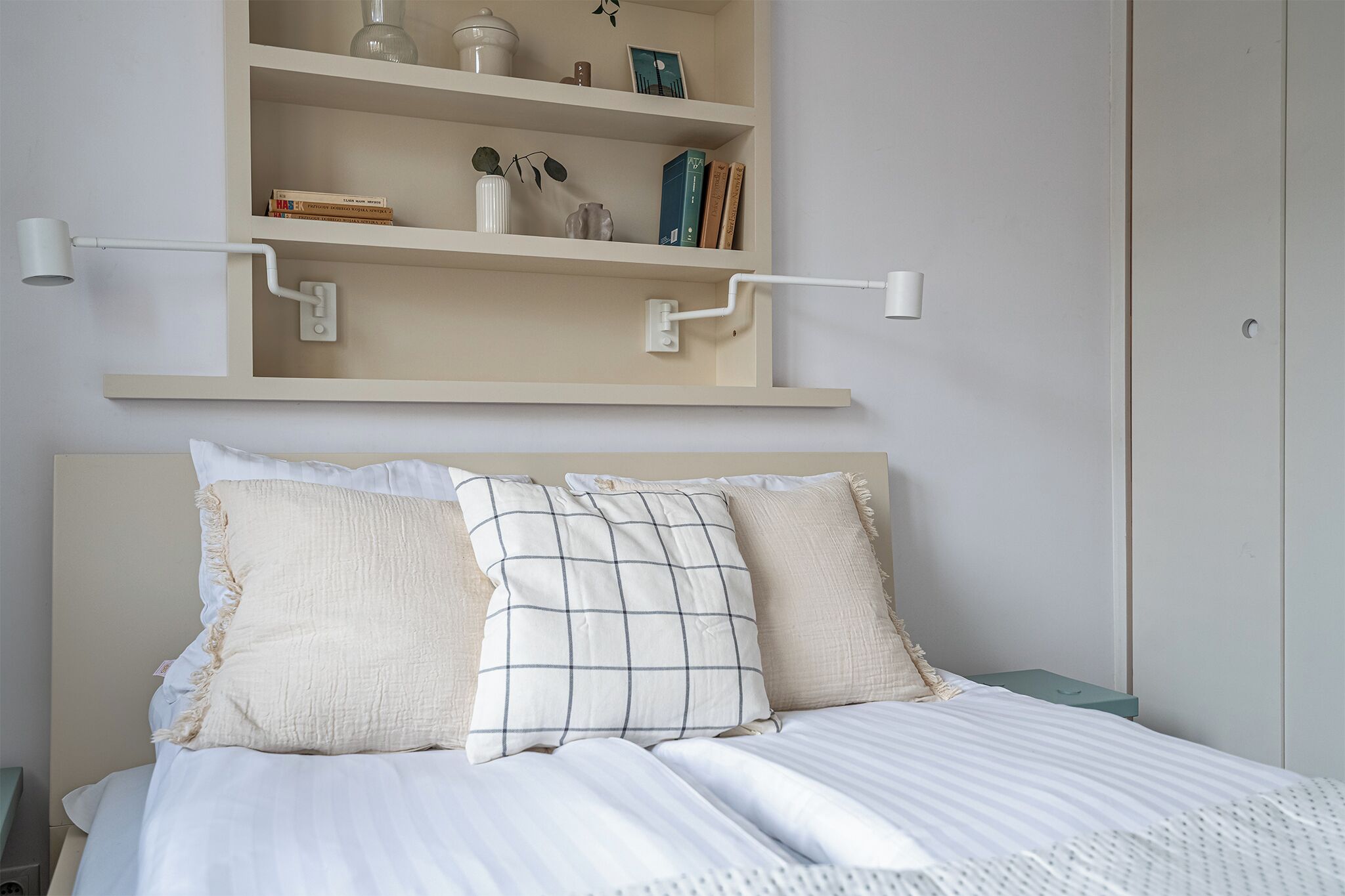 A neatly made bed with a headboard, a shelf above with books and decorative items, and a wall-mounted light fixture.