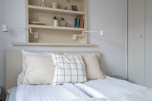 A neatly made bed with a headboard, a shelf above with books and decorative items, and a wall-mounted light fixture. - RentPlanet - Aleje Jerozolimskie II (Warsaw)
