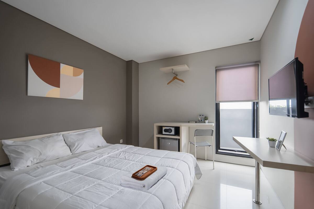Ein modernes Hotelzimmer mit Bett, Schreibtisch, Stuhl, Fernseher und einem Fenster mit Jalousien.