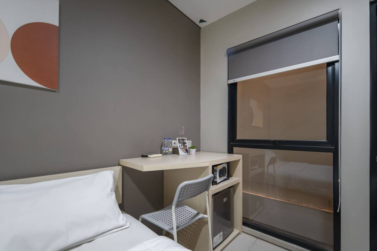 Ein kleines Hotelzimmer mit Bett, Schreibtisch, Stuhl, einem kleinen Schrank und einem Fenster mit Aussicht.
