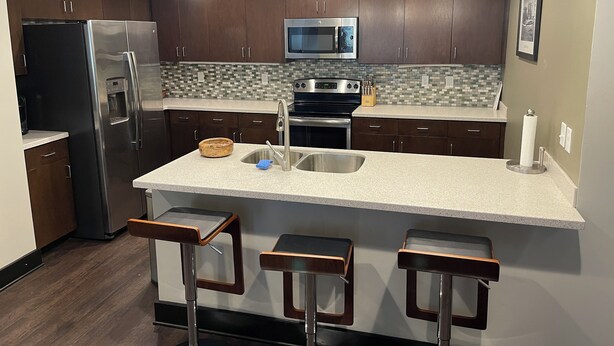 A modern kitchen with dark wood cabinets, a stainless steel refrigerator, a microwave, and a large island with a sink. There are three bar stools in front of the island. - Beautiful Downtown Condo! Steps away from the best dining in town! (Tallahassee)