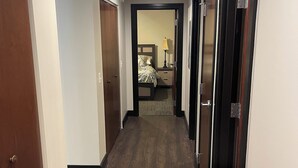 A hotel hallway with wooden flooring, a bed with a nightstand and lamp, and a door at the end. - Beautiful Downtown Condo! Steps away from the best dining in town! (Tallahassee)