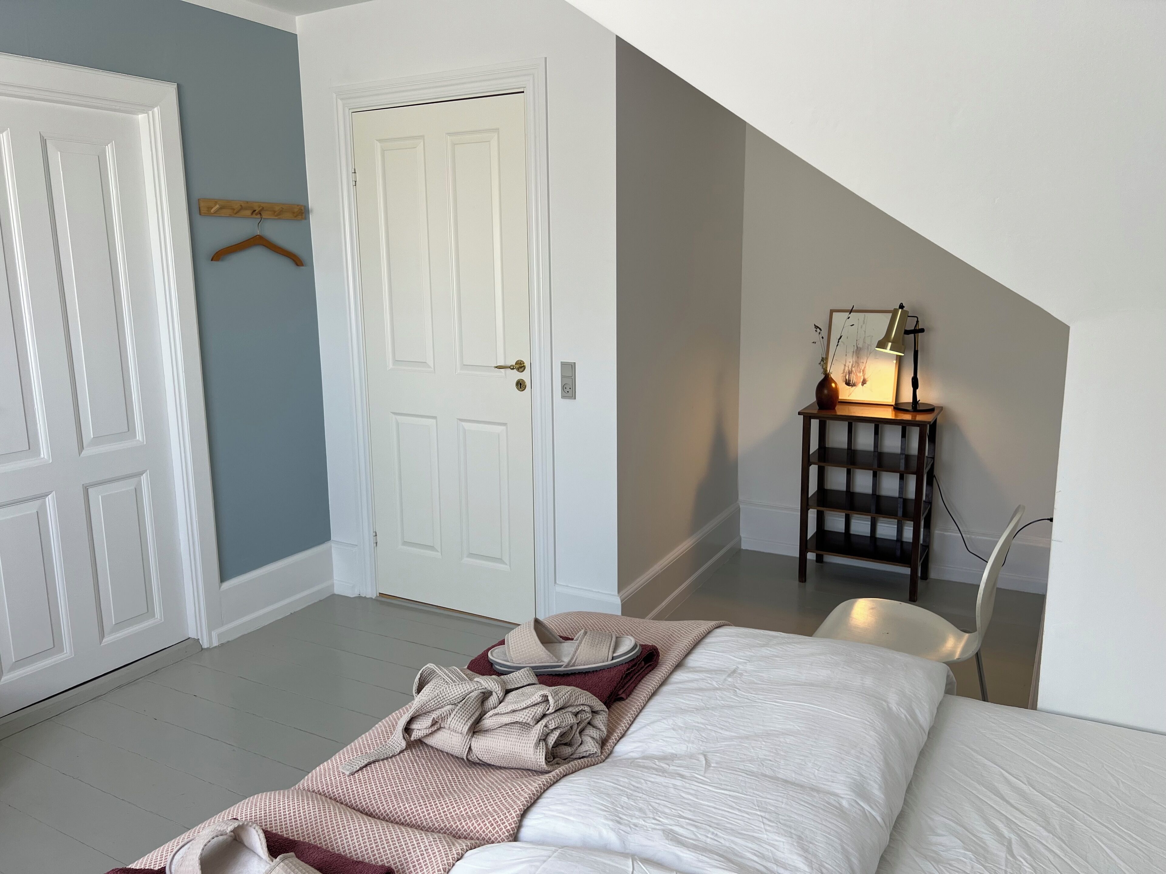 A bedroom with a bed, a chair, a desk with a lamp, and a door.