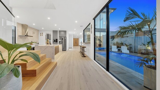 A modern kitchen with a central island, a dining area with bar stools, and a large sliding glass door leading to a pool area with lounge chairs. - Luxe designer house with Pool, Cinema & Gym (Clyde North)