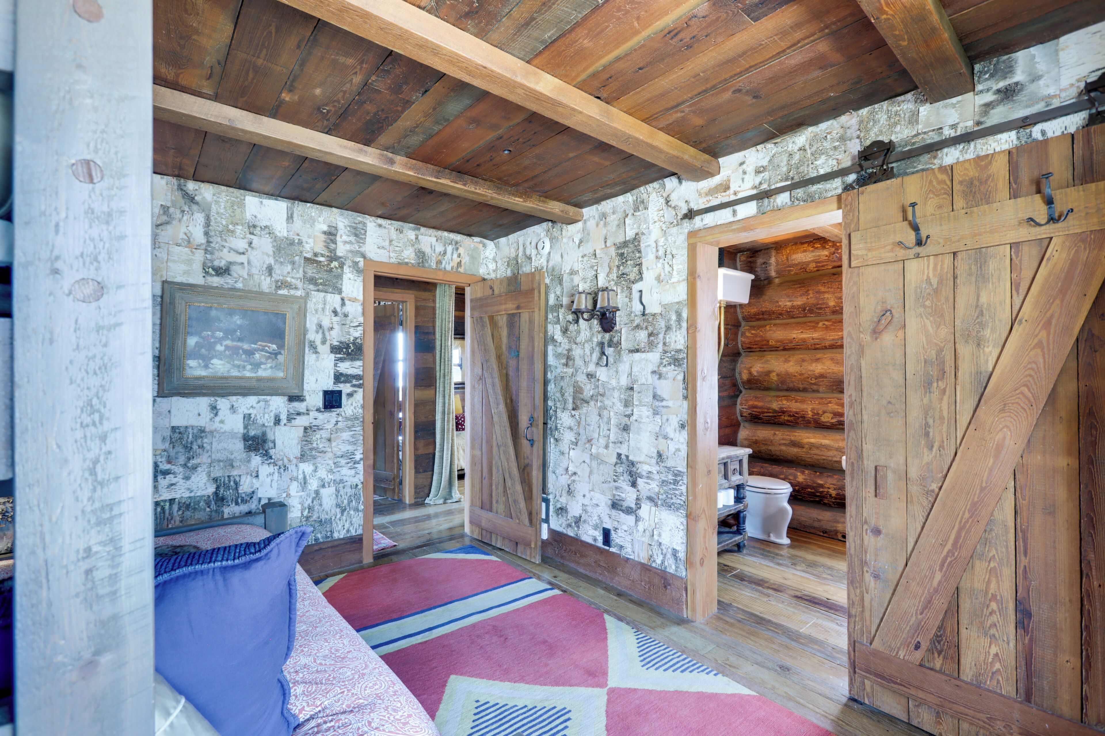 A rustic log cabin interior with a wooden ceiling, stone walls, a bed with a patterned bedspread, and a wooden door.