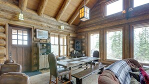 A log cabin interior with a dining table, chairs, a sofa, and large windows offering a view of the forest.