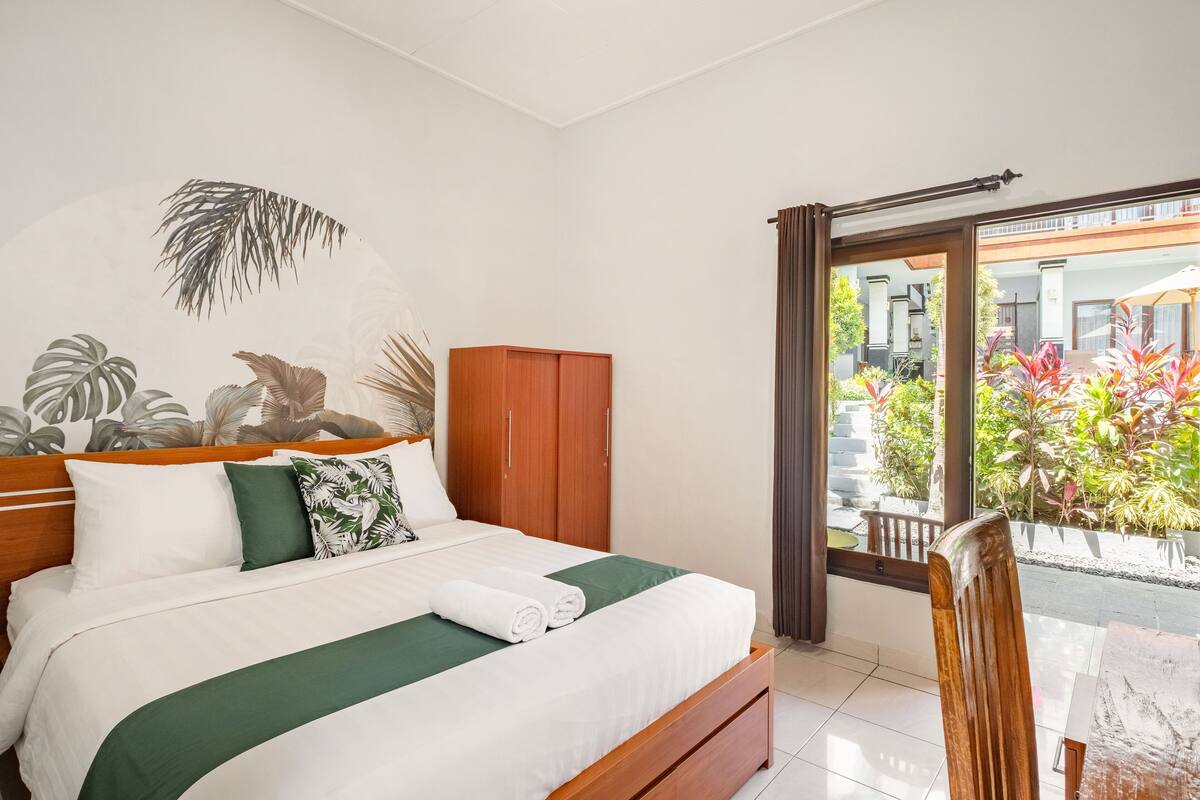 A hotel room with a bed, wooden wardrobe, and a view of a garden through a sliding door.