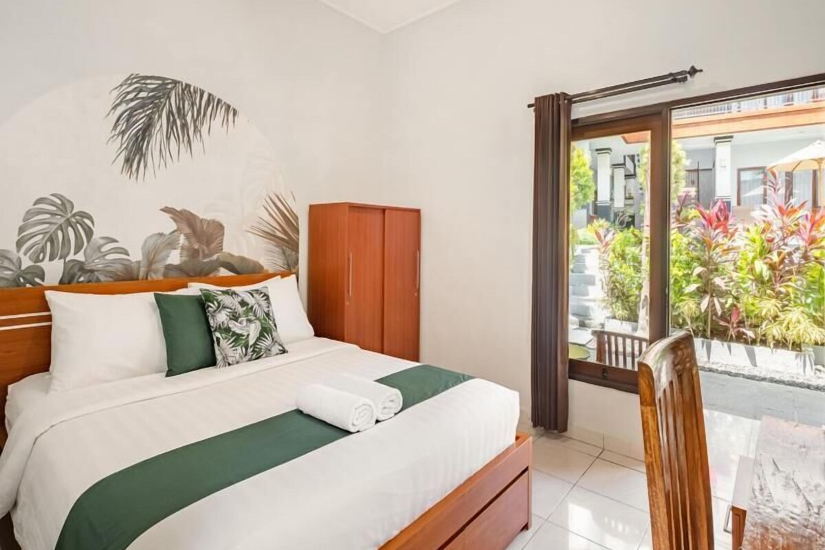 A hotel room with a bed, a wooden wardrobe, a green bedspread, and a view of a garden through a sliding door.