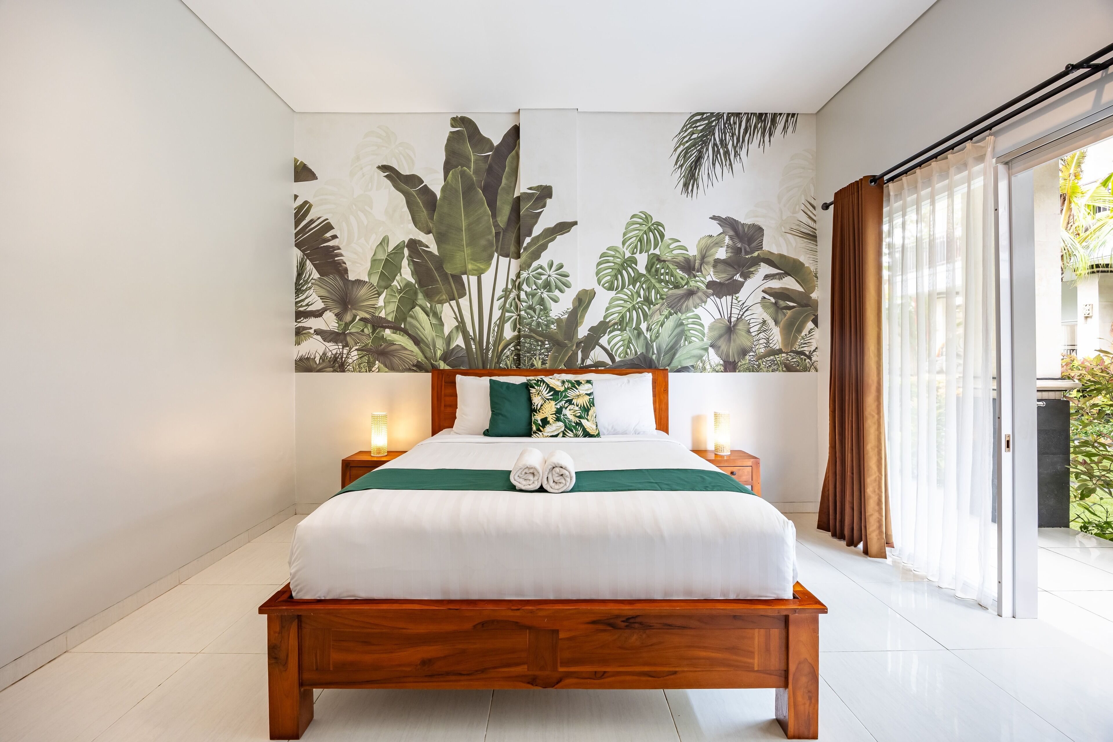 A bedroom with a wooden bed, white bedding, a tropical plant mural, and a sliding glass door to a balcony.