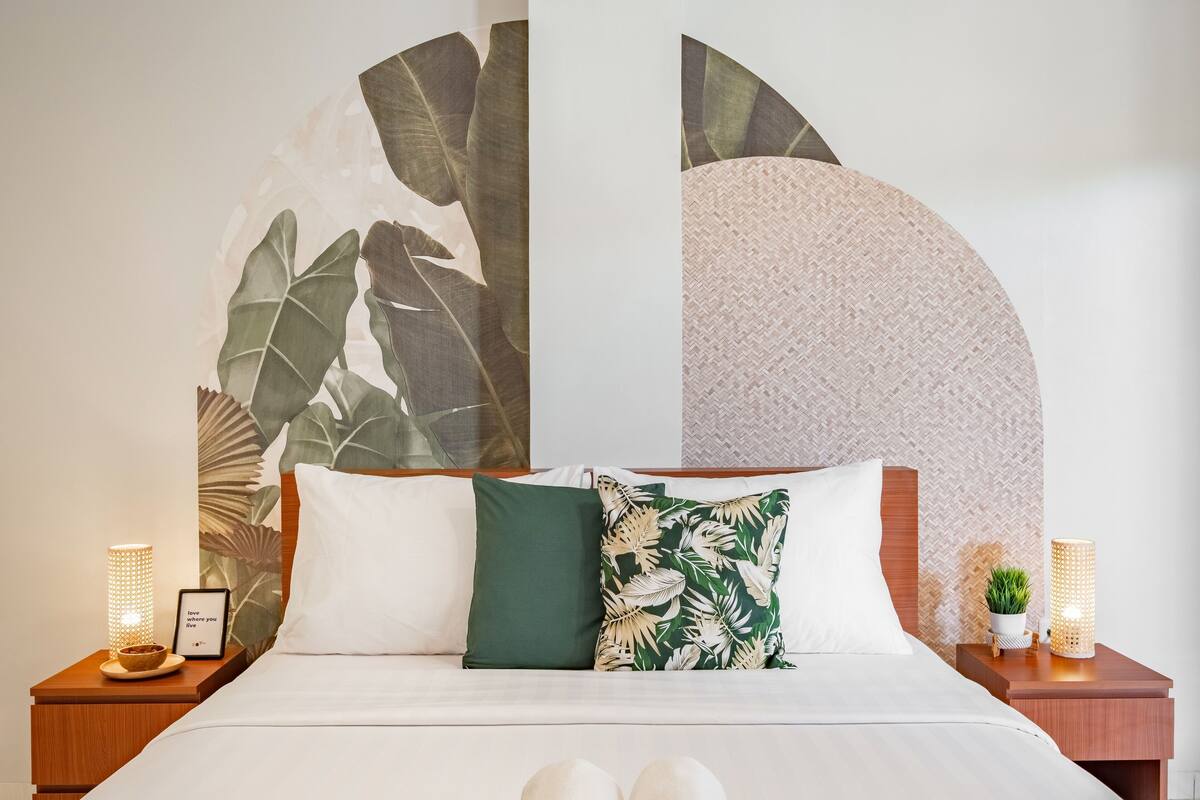 A neatly made bed with a white comforter and two pillows, one green and one with a tropical leaf pattern. There are two bedside tables with lamps and a framed picture on one.