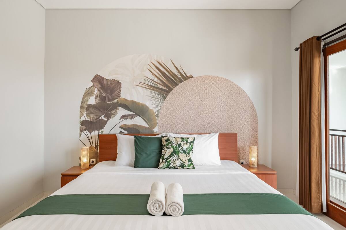 A neatly made bed with rolled towels, a green and white bedspread, and a tropical leaf wall art.