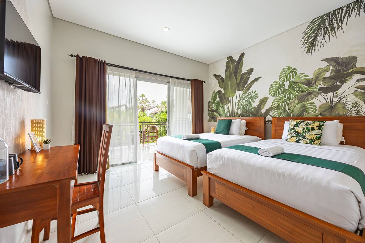 A hotel room with two beds, a wooden desk, a chair, a TV, and a balcony with a view of greenery.