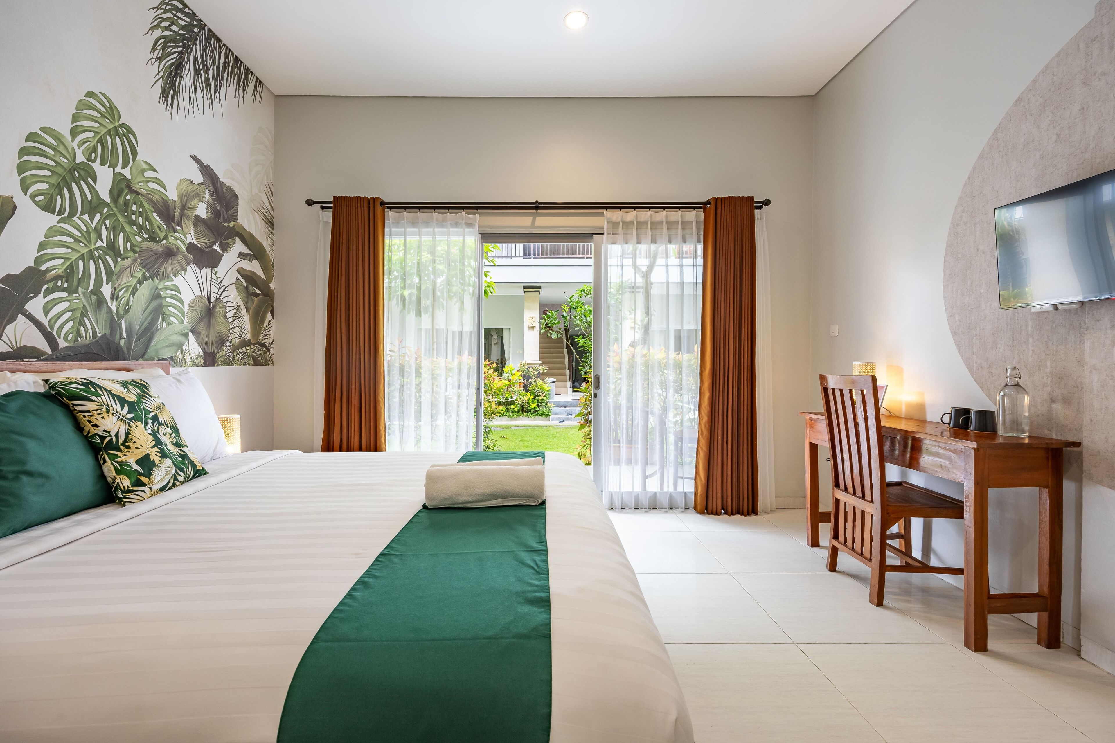 Une chambre d’hôtel avec un grand lit, un bureau avec une chaise, un téléviseur fixé au mur et une vue sur le jardin à travers les rideaux ouverts.