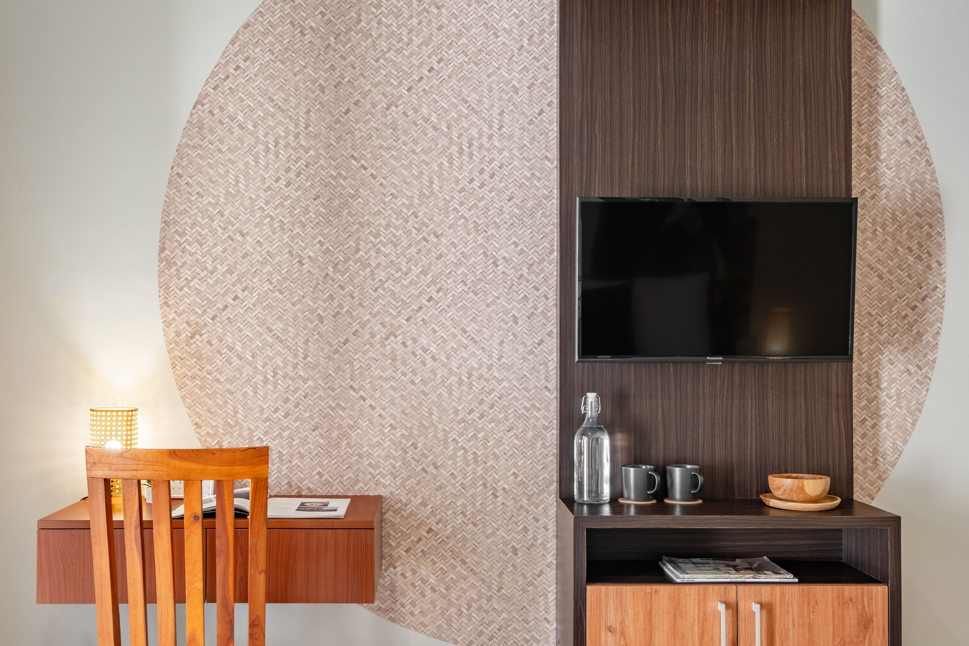 A modern room with a large woven wall, a wooden chair, a small table with a lamp, a TV on a wooden shelf, and a cabinet with a glass bottle and cups.