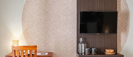 A modern room with a large woven wall, a wooden chair, a small table with a lamp, a TV on a wooden shelf, and a cabinet with a glass bottle and cups.