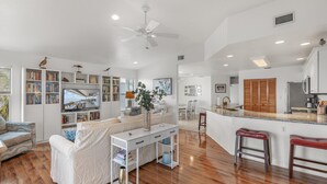 A spacious living area with a large white sofa, a coffee table, and a bookshelf. There is a kitchen area with a refrigerator, microwave, and wooden cabinets. - Fenimore Mill 6DE Dual Delight, Two Units in One - Families/Groups/Gulf Front! (Cedar Key)