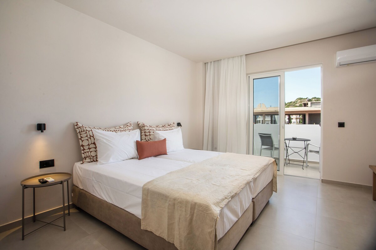A modern hotel room with a large bed, a small side table, and a view of the outside balcony.