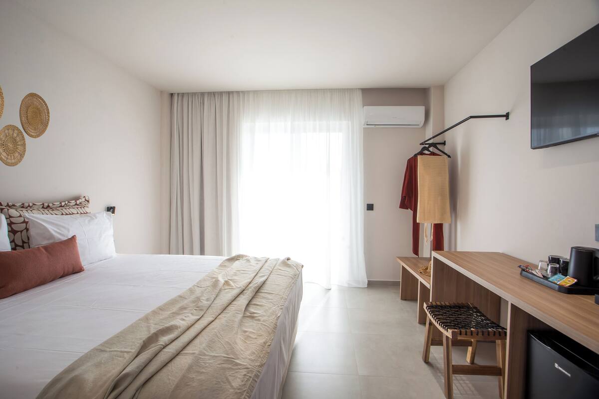 A modern hotel room with a large bed, a desk with a chair, a TV mounted on the wall, and a window with curtains.