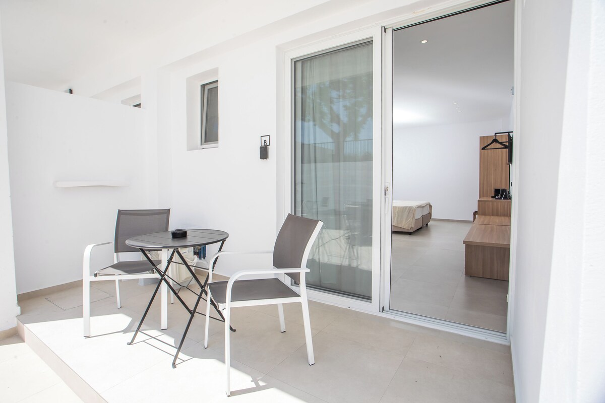 A modern outdoor patio with a table and chairs, a sliding glass door leading to a balcony, and a minimalist interior visible through the door.