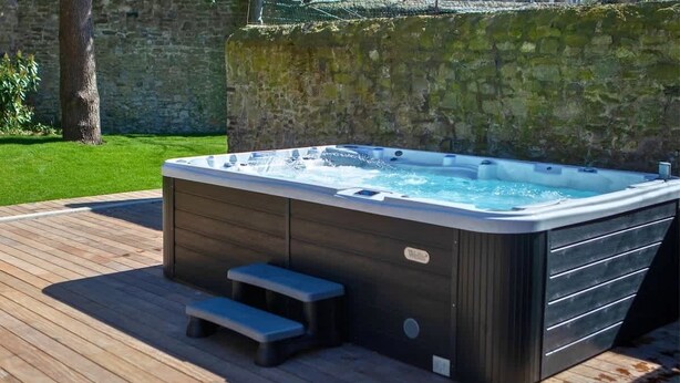 A hot tub on a wooden deck with steps leading up to it, surrounded by a stone wall and greenery.