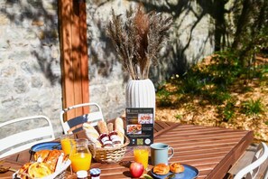 Ein Frühstückstisch mit Brot, Gebäck, Früchten und Saft vor einer Steinwand mit Grünpflanzen.
