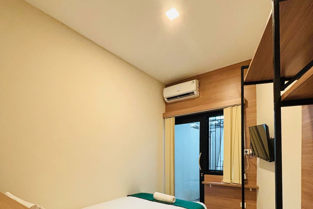 A hotel room with a single bed, a desk with a chair, a TV, and an air conditioning unit on the wall.