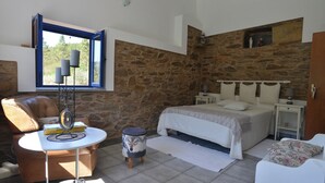 Een slaapkamer met een stenen muur, een bed met witte beddengoed, een klein rond tafeltje met een decoratief object, een stoel en een raam met gordijnen.