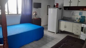 Ein Zimmer mit einem Bett, einer kleinen Küchenzeile, einem Fernseher und einem fenster mit Vorhängen.
