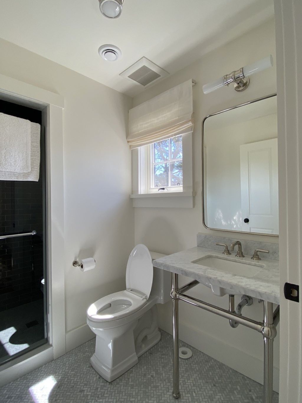 A bathroom with a toilet, sink, mirror, window with blinds, and a shower area.