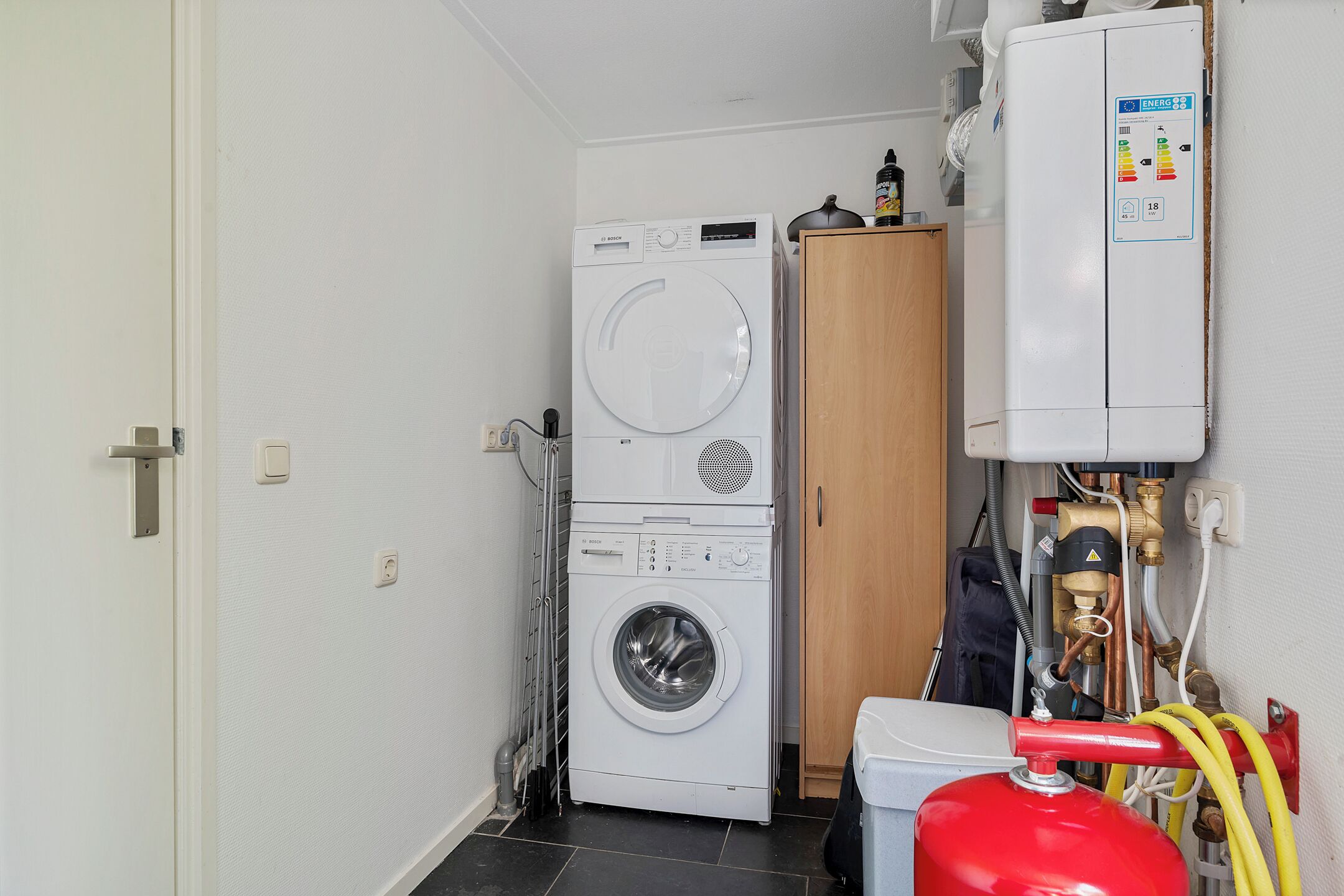 Une petite buanderie équipée d’un lave-linge et d’un sèche-linge superposés, d’un meuble en bois et dont la tuyauterie est visible.