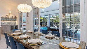 A modern dining area with a glass table set for six, blue chairs, and a large window overlooking a pool. - Relaxing 3BR / 2BA Naples Retreat - w Private Heated Pool (Naples)