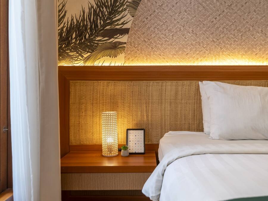 Cama bem arrumada com roupa de cama branca e um travesseiro verde, uma pequena mesa de cabeceira com uma luminária e um quadro emoldurado, e uma parede texturizada atrás da cama.