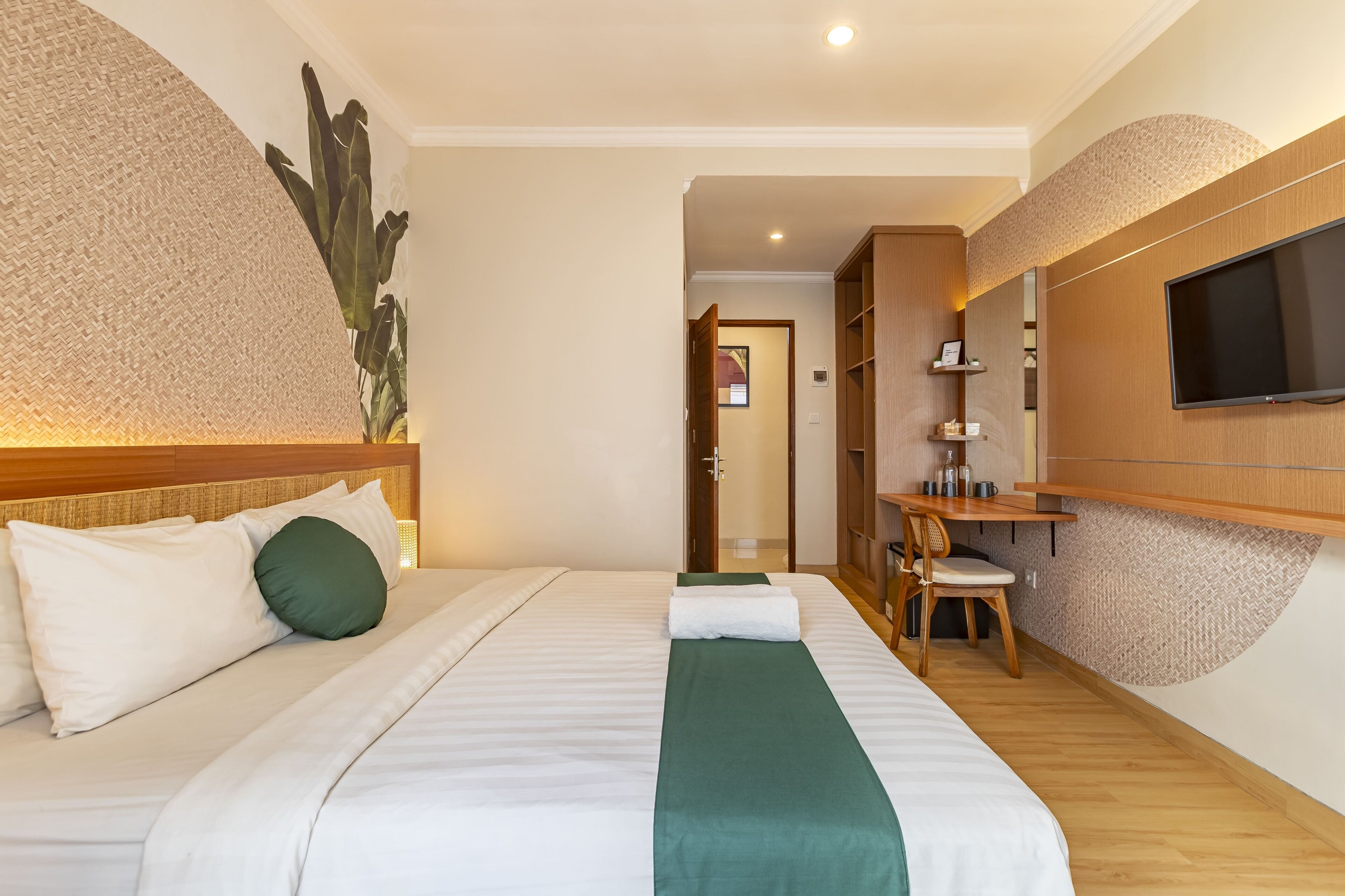 A hotel room with a bed, a desk, a chair, a TV, and a large plant.