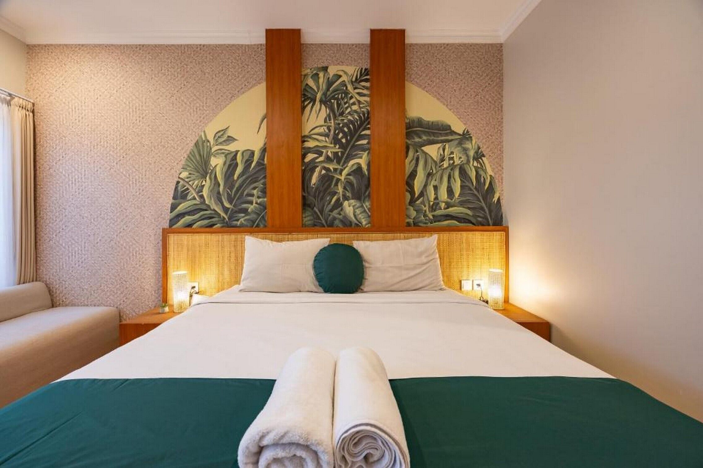 A hotel room with a large bed, a green bedspread, two rolled towels, a green pillow, and a wall mural of tropical plants.