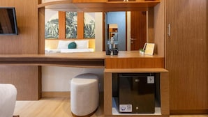 Camera d'albergo compatta con una scrivania in legno, un piccolo frigorifero, uno sgabello imbottito e un letto con cuscino verde.