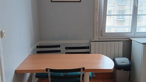 Una habitación con una mesa y sillas de madera, un radiador, una ventana con vistas a edificios y un cuadro en la pared.