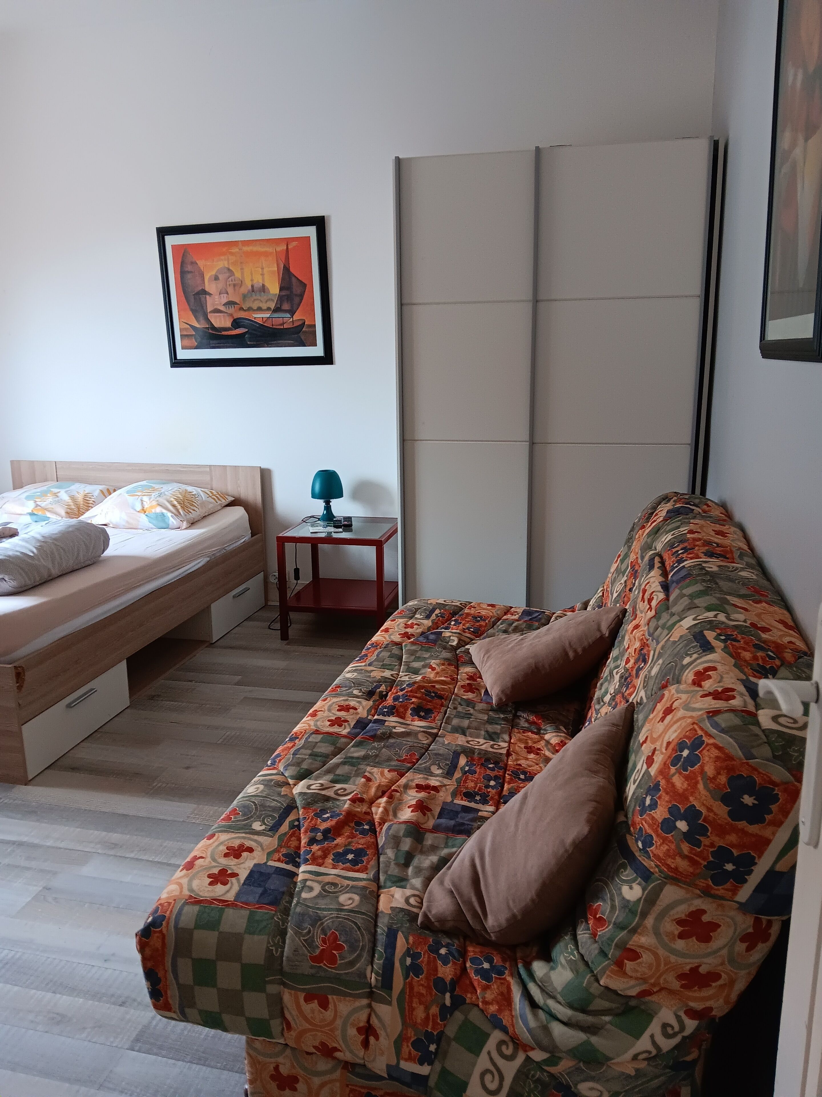 Un dormitorio con cama, sofá, una mesita de noche con lámpara y un cuadro en la pared.