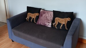 Un sofá azul con cojines de estampado de leopardo y una almohada con estampado de león, colocado frente a una pared azul claro con dos cuadros enmarcados.