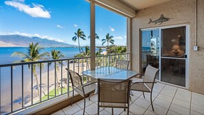 A balcony with a glass table and chairs, overlooking a beach with palm trees and mountains. - Menehune Shores, #609 2 Bedroom Condo (Kihei)