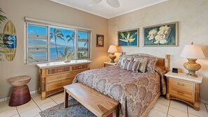 A bedroom with a bed, a nightstand, a lamp, a bench, a window with a view, and a ceiling fan. - Menehune Shores, #609 2 Bedroom Condo (Kihei)