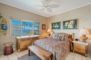 A bedroom with a bed, a nightstand, a lamp, a bench, a window with a view, and a ceiling fan.