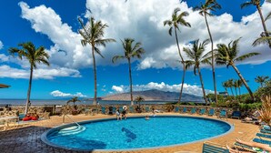 A swimming pool with lounge chairs and palm trees, surrounded by a stone wall and a view of the ocean and mountains. - Menehune Shores, #609 2 Bedroom Condo (Kihei)