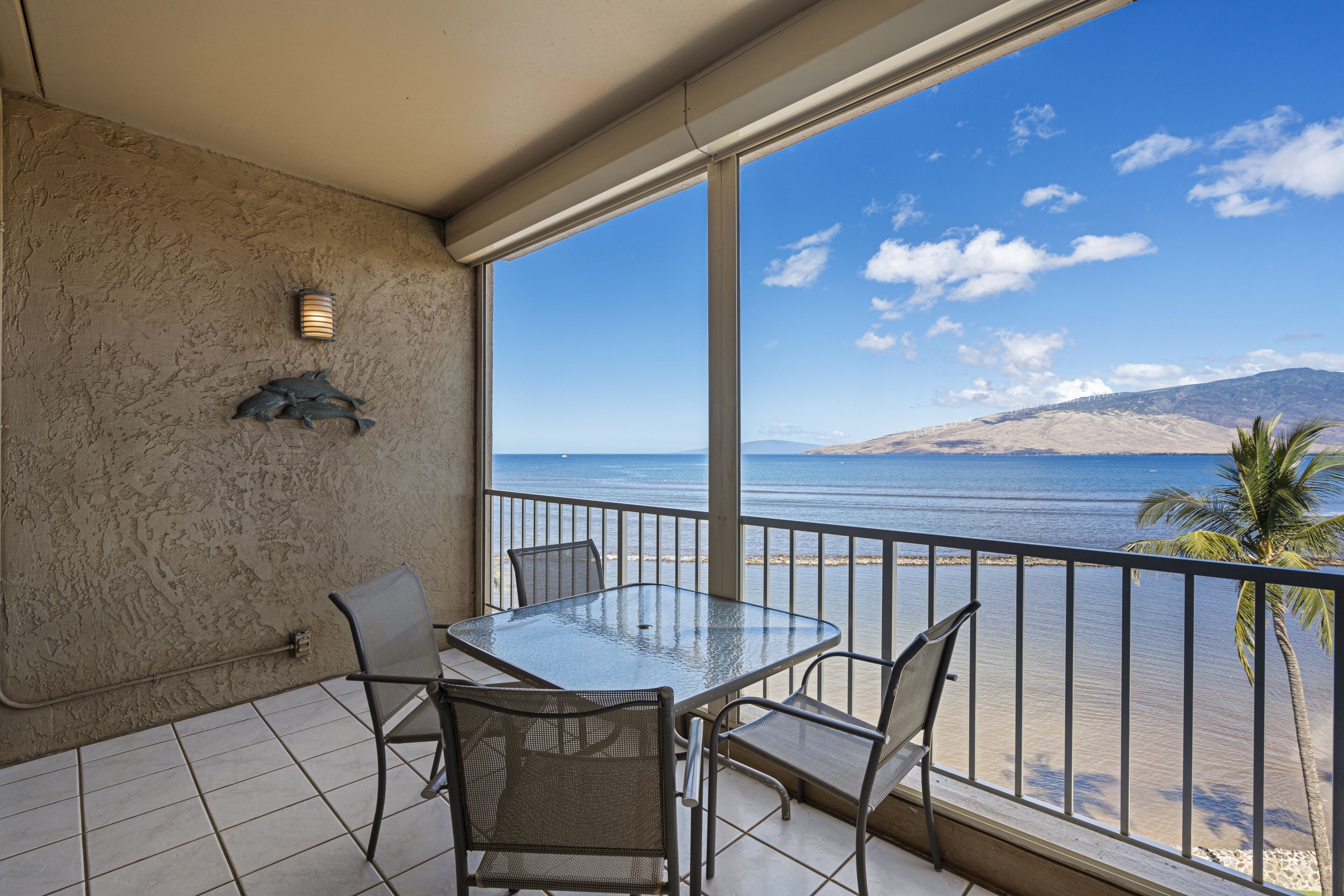 A balcony with a glass table and chairs, a wall-mounted light fixture, and a view of the sea and a mountain.