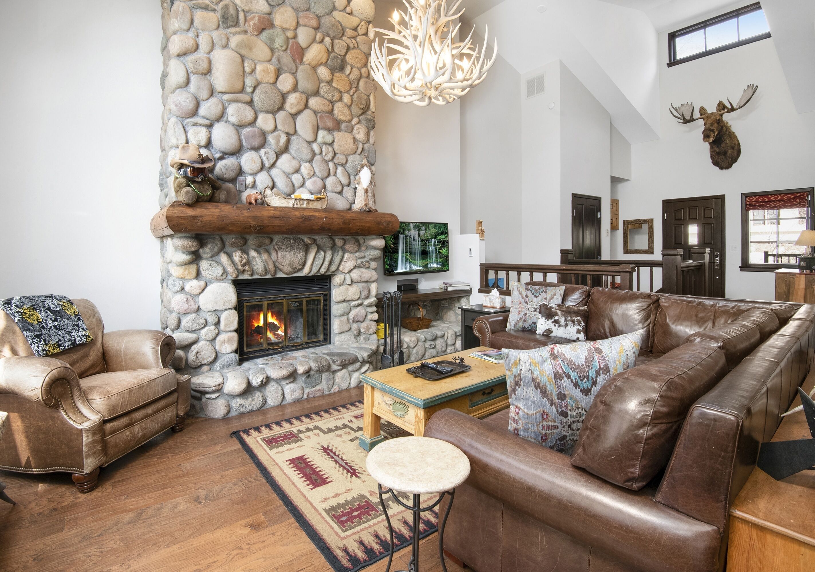 Una sala de estar acogedora con chimenea de piedra, muebles de cuero y un ciervo colgado en la pared.