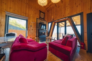 A wooden cabin interior with a fireplace, red sofas, a dining table, and large windows offering a view of the outdoors.